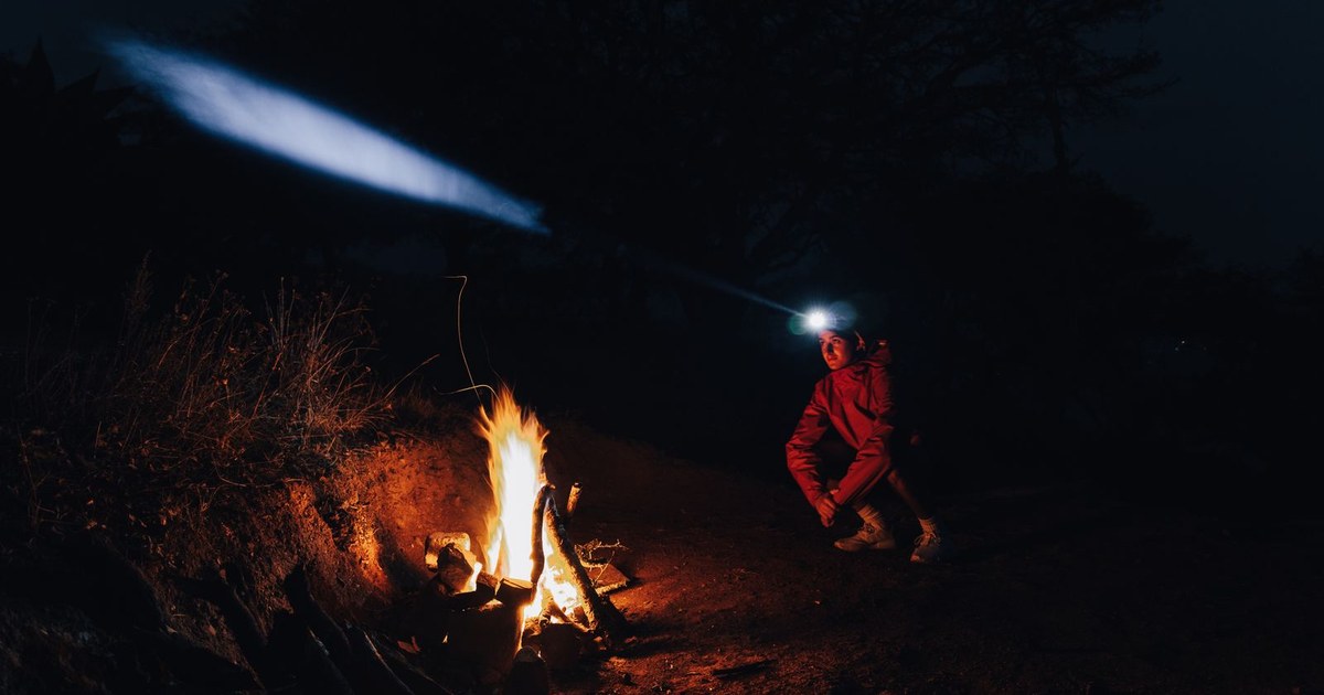 Man with headlamp sitting by campfire at night, surrounded by nature.