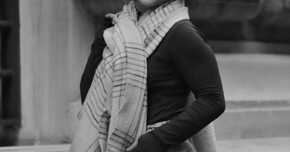 Black and white portrait of a smiling woman in urban setting, wearing scarf and casual attire.