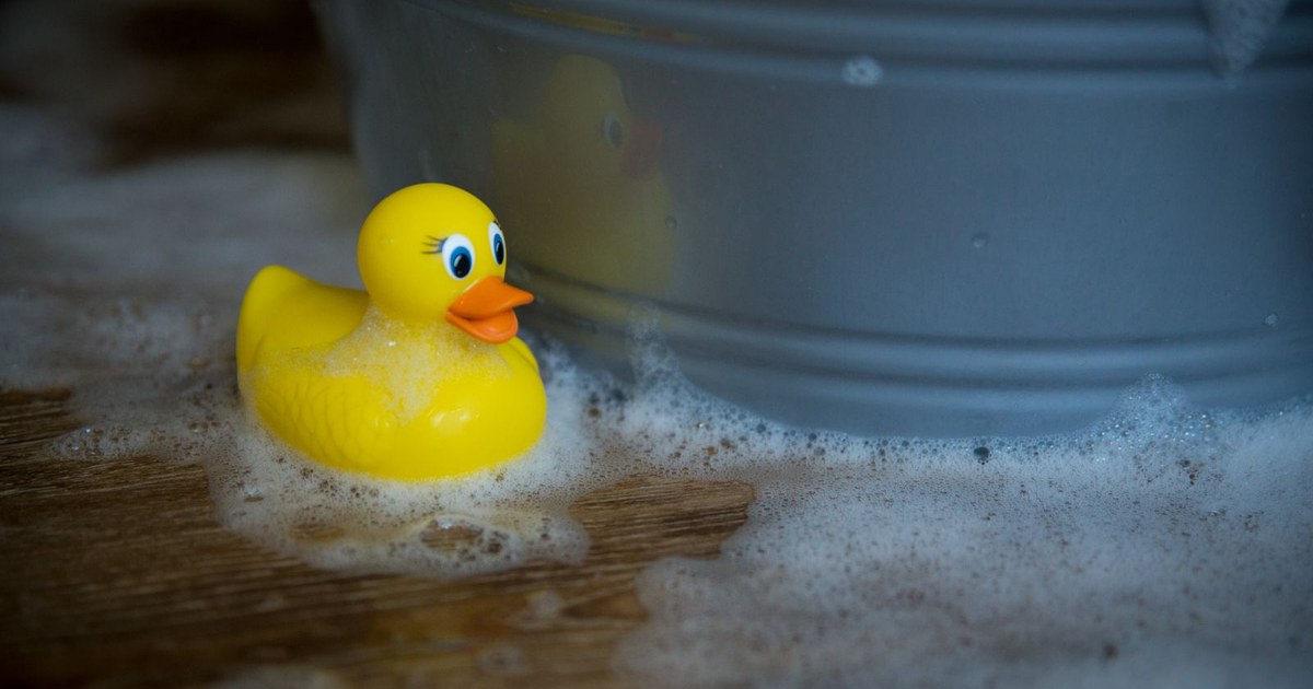A yellow rubber duck floating near soap bubbles beside a tub on a wooden surface.