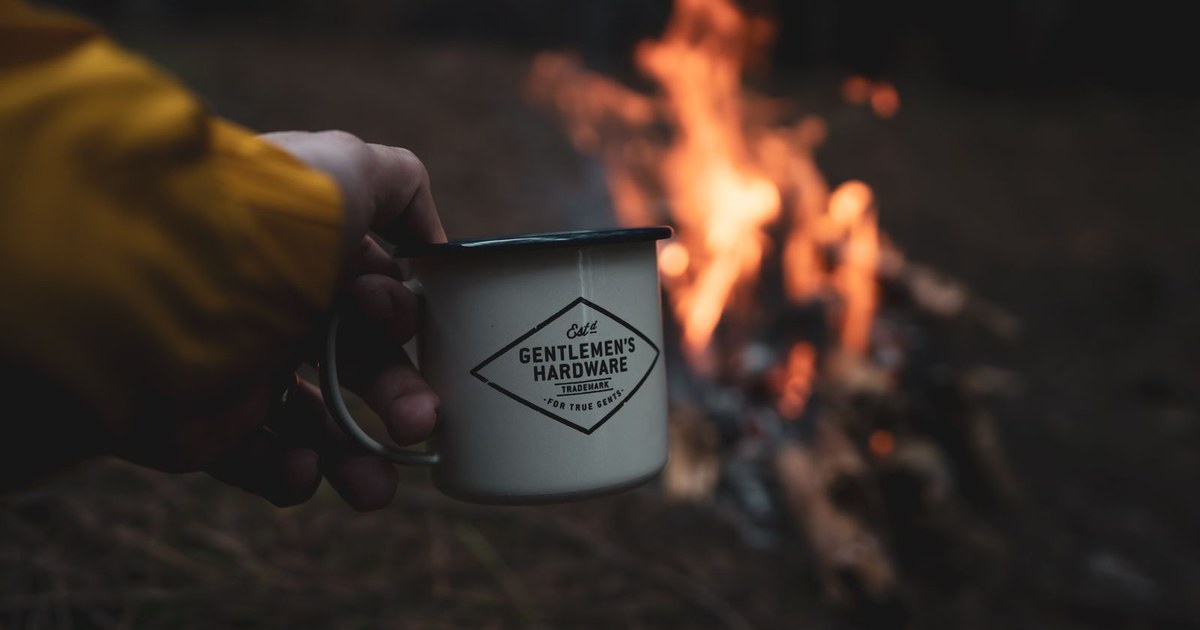A hand holding a mug in front of a warm campfire, creating a cozy outdoor vibe.
