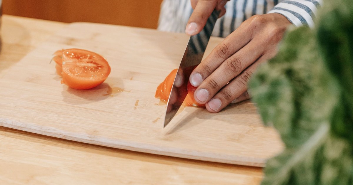 Crop anonymous ethnic male in striped shirt cutting ripe tomato with knife on chopping board in kitchen