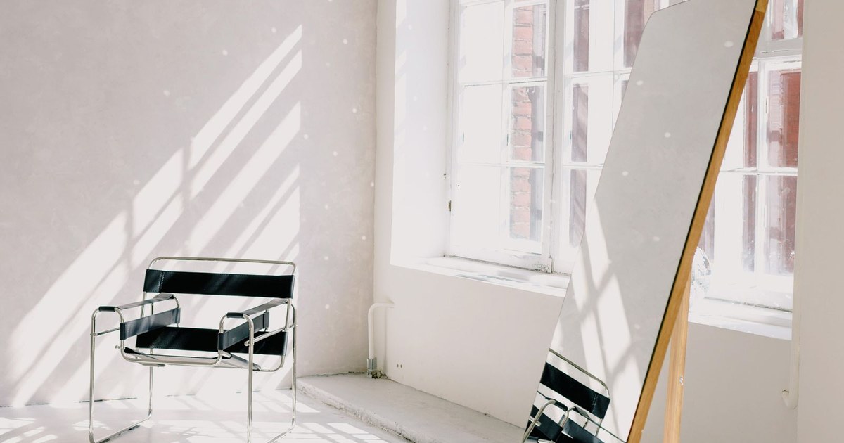 Bright minimalist room with a modern armchair, large mirror, and sunlight casting dramatic shadows.