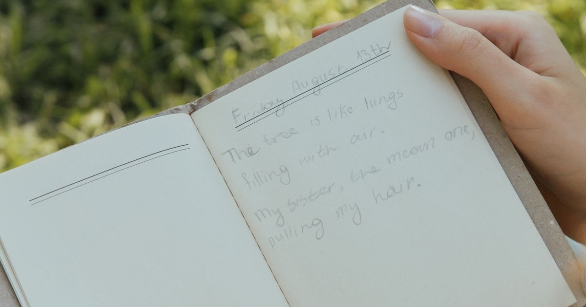 A woman writes in a journal outdoors on a sunny day, surrounded by grass.