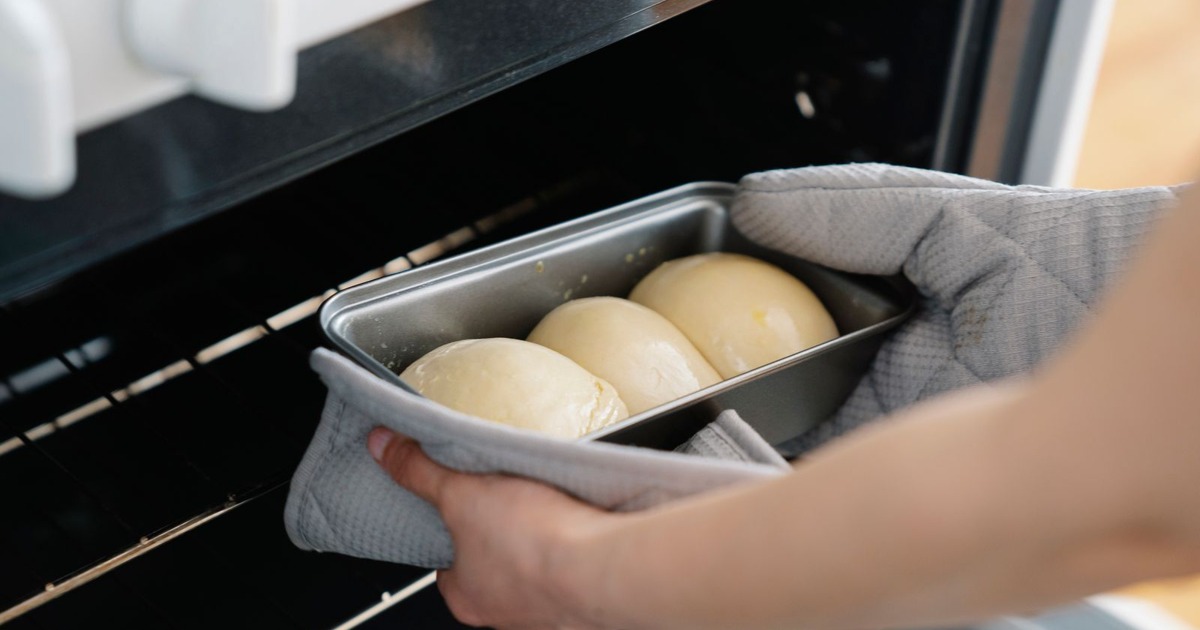 Person placing dough buns in oven, wearing gloves and using baking tray. Captured mid-preparation in a kitchen setting.