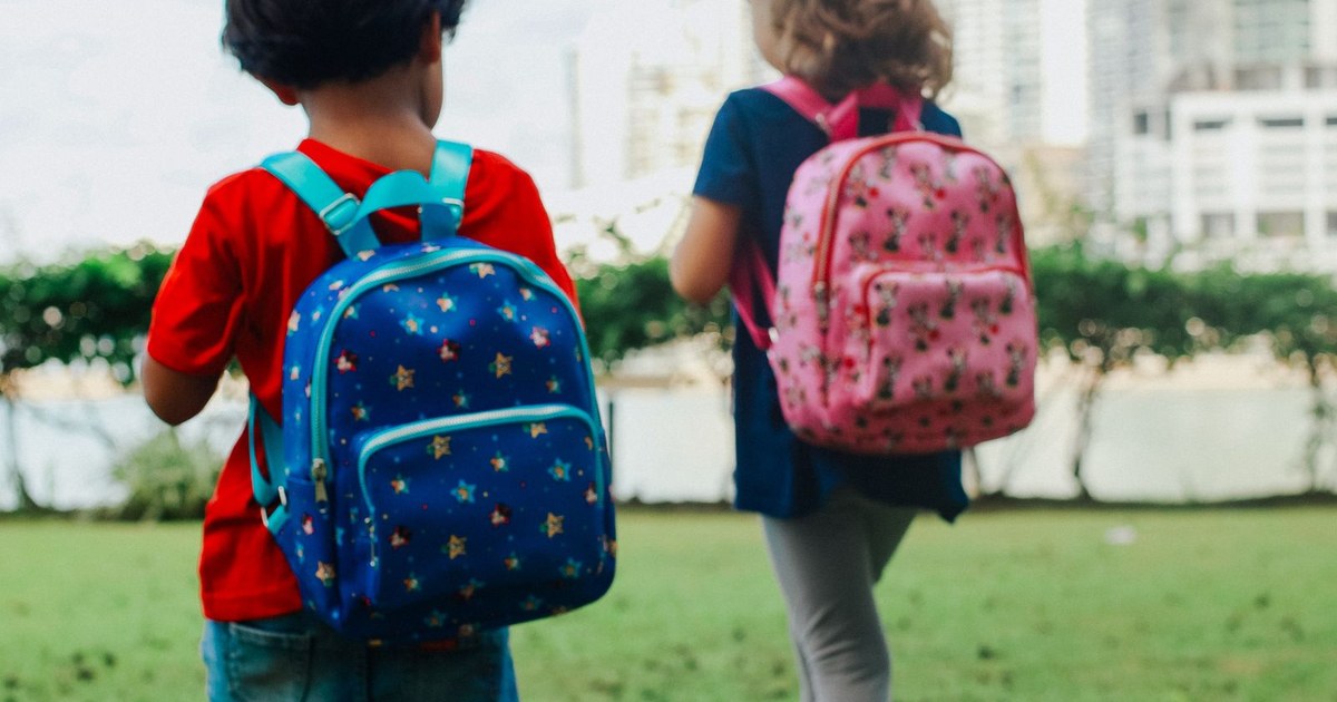 Two kids with colorful backpacks strolling through a city's green park in daylight.