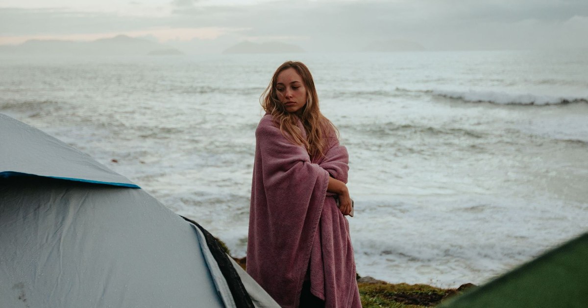 Woman wrapped in blanket on a coastal camping trip at sunset, near ocean waves.
