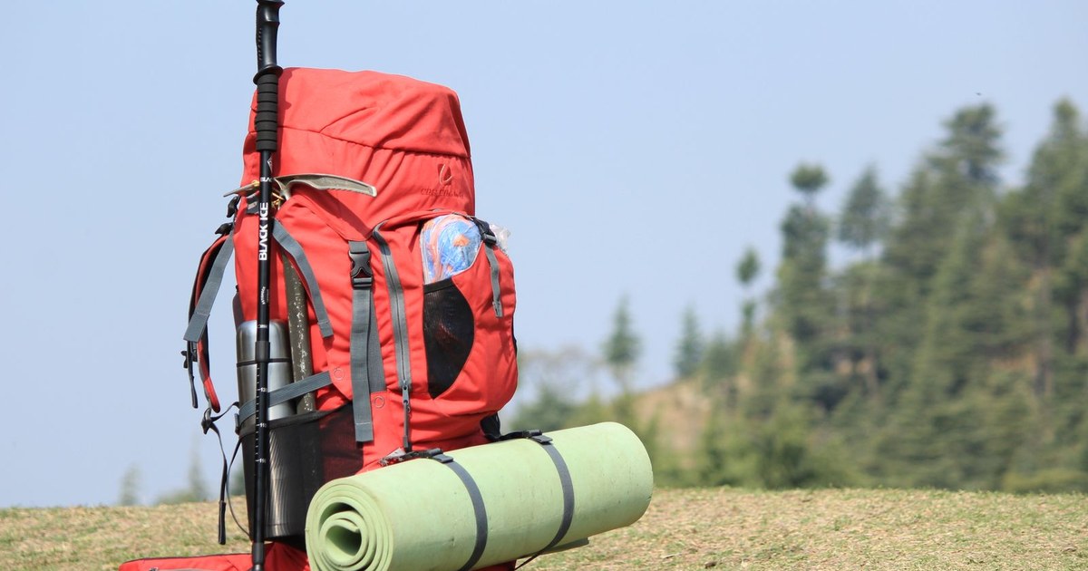 Red backpack with hiking pole and mat on grassy field with forest background.